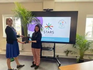 Helen, CEO, accepts plants from Katie from Poundbury Gardens in front of a large TV screen with the STARS and Interstellar logos visible.